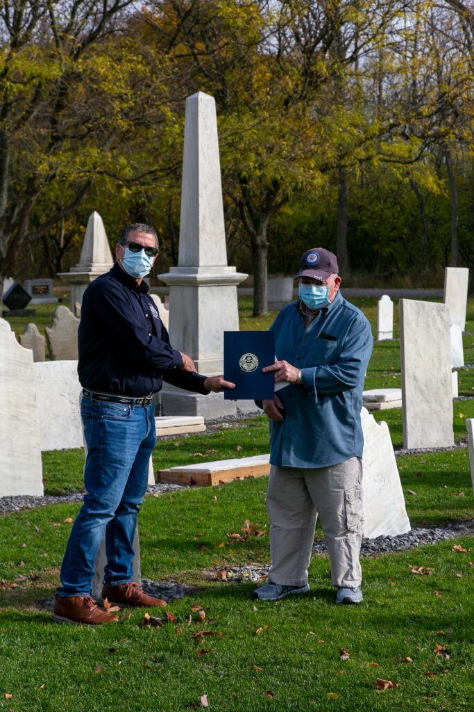 Two men standing in a cemetary holding a plaque