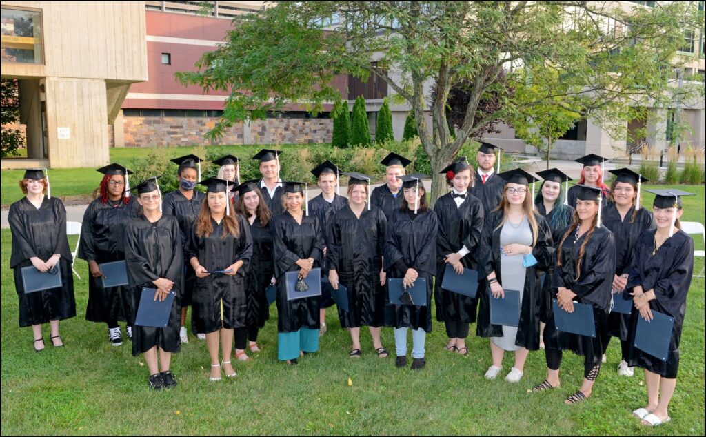 About 24 graduates standing outside under a tree