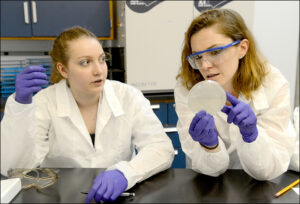 Jessica Halliley, right, holding a petri dish and talking to a student in a lab